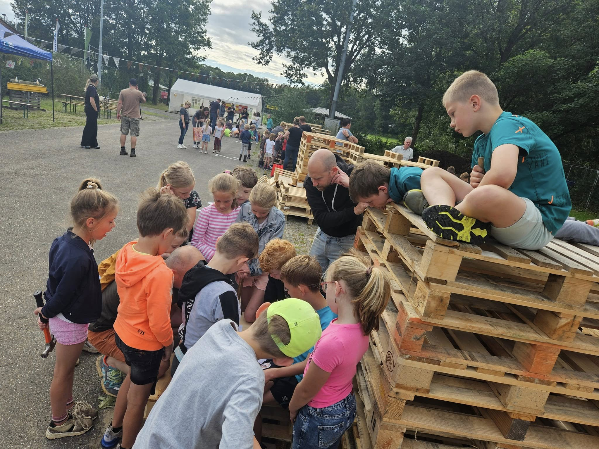 Allemaal kinderen die aandachtig naar iets aan het kijken zijn. Één van de kinderen zit op een hoge stapel pallets, en één kind ligt op de hoge stapel pallets
