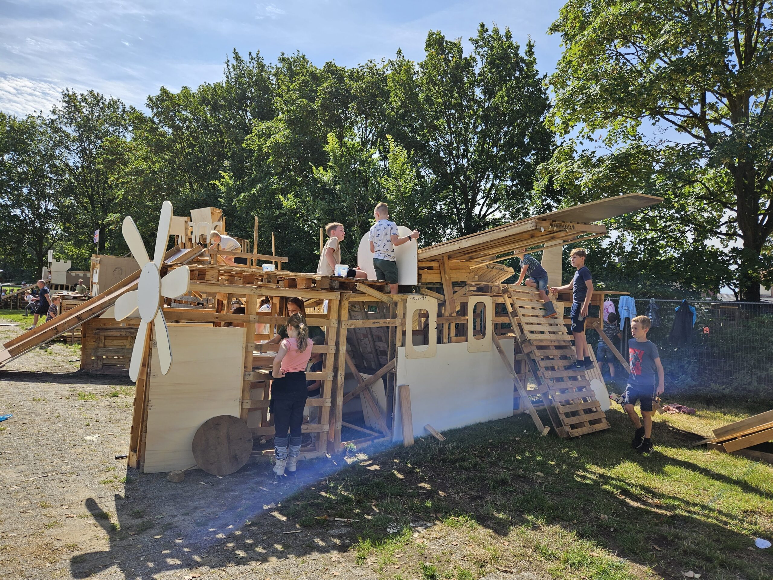 Een groot kunstwerk met een trap gemaakt van pallets, een houten zonnenbloem en een grote plank die naast het kunstwerk uitsteekt
