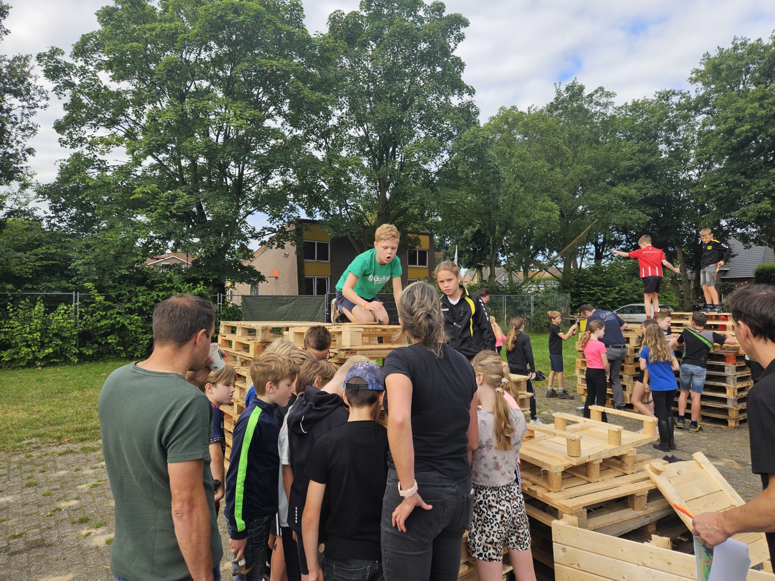 Een hoop kinderen en een drietal begeleiders bij aantal stapels pallets, een van de kinderen zit er op en in de achtergrond kan je zien dat er twee jongens op staan